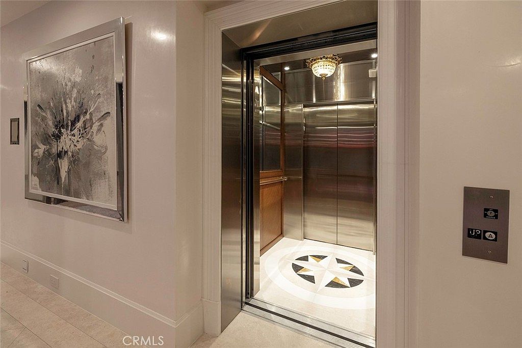 The image showcases an interior elevator with stainless steel doors and a decorative floor pattern. The elevator's interior is illuminated by a chandelier, adding a touch of elegance. The adjacent wall features elevator control buttons and a framed artwork, contributing to a sophisticated and functional design.