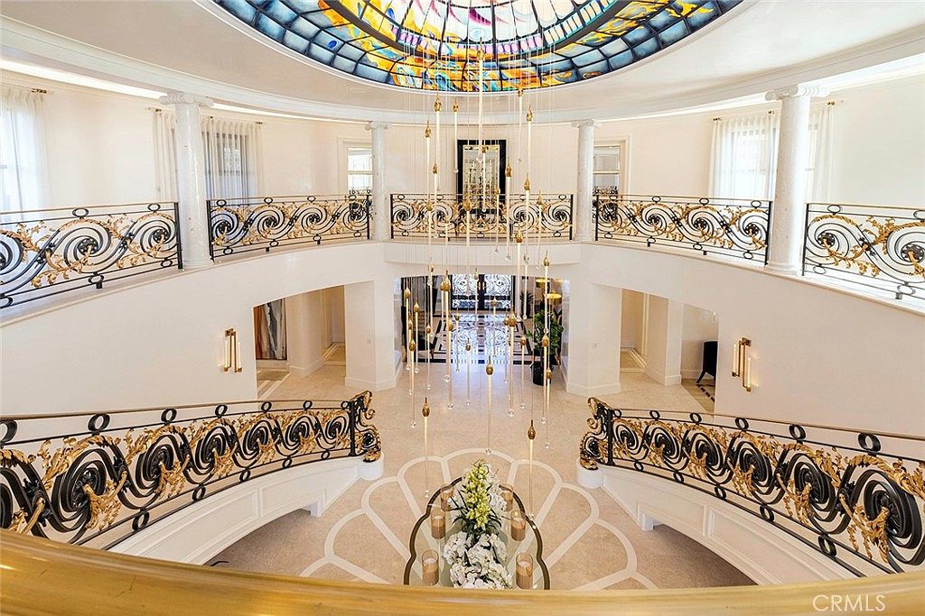 This grand foyer features a stunning stained-glass dome ceiling and an ornate wrought-iron railing that overlooks the lower level. A cascading chandelier adds to the elegance, while the marble flooring and decorative accents create a luxurious and inviting atmosphere. The view is from the upper level, looking down at the foyer.