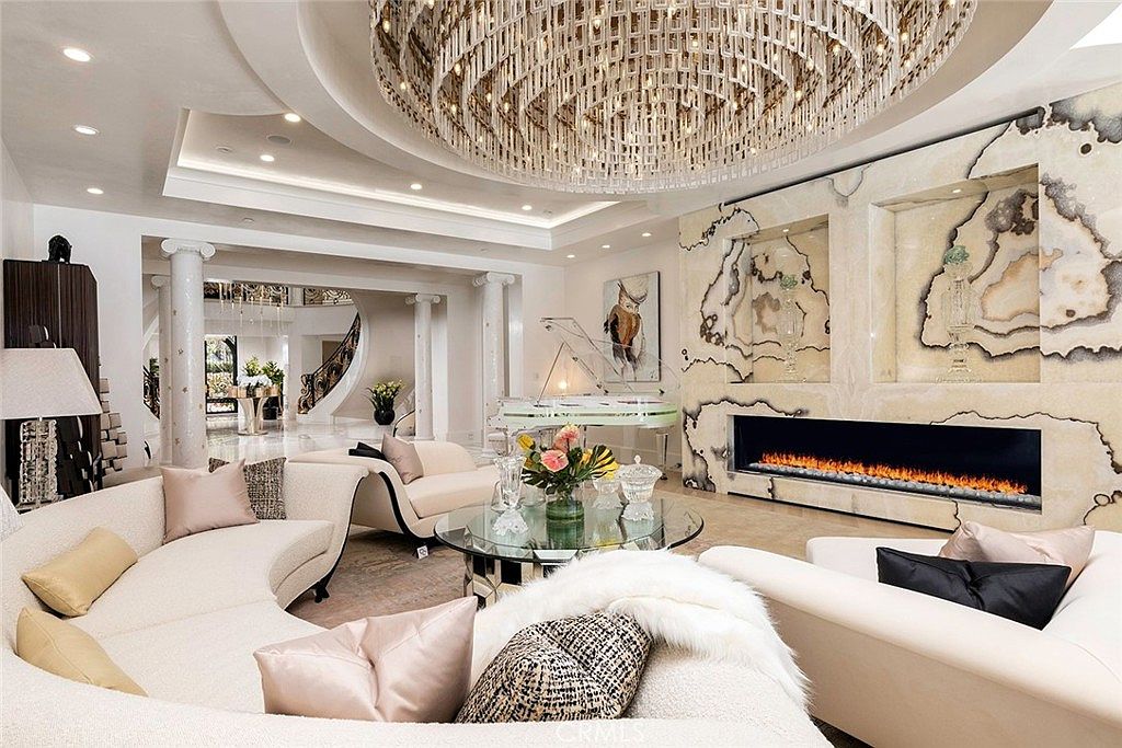 This grand living room features a luxurious and opulent design with a massive chandelier as a focal point. The room includes curved sofas, a glass coffee table, a modern fireplace, and a clear grand piano, all set against a backdrop of marble-like wall panels. The space is open and airy, with columns leading to a staircase, creating an impressive and elegant atmosphere.