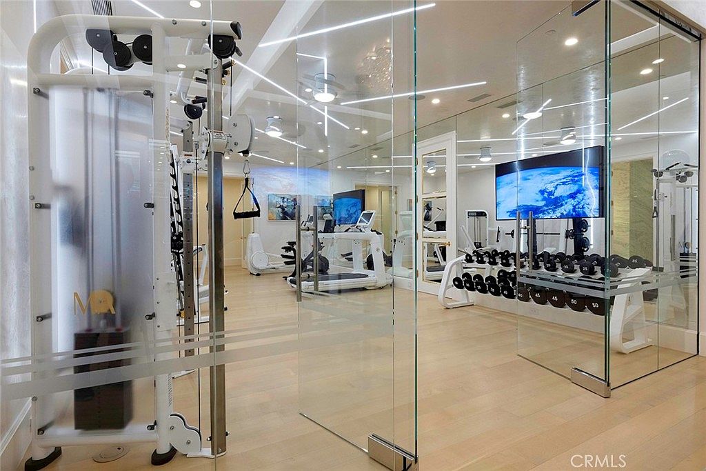 This is a modern home gym featuring glass walls, light wood flooring, and recessed lighting. The gym is equipped with various exercise machines, including a weight training system, treadmills, and a dumbbell rack with a television mounted on the wall. The space appears clean, well-lit, and designed for a comprehensive workout experience.