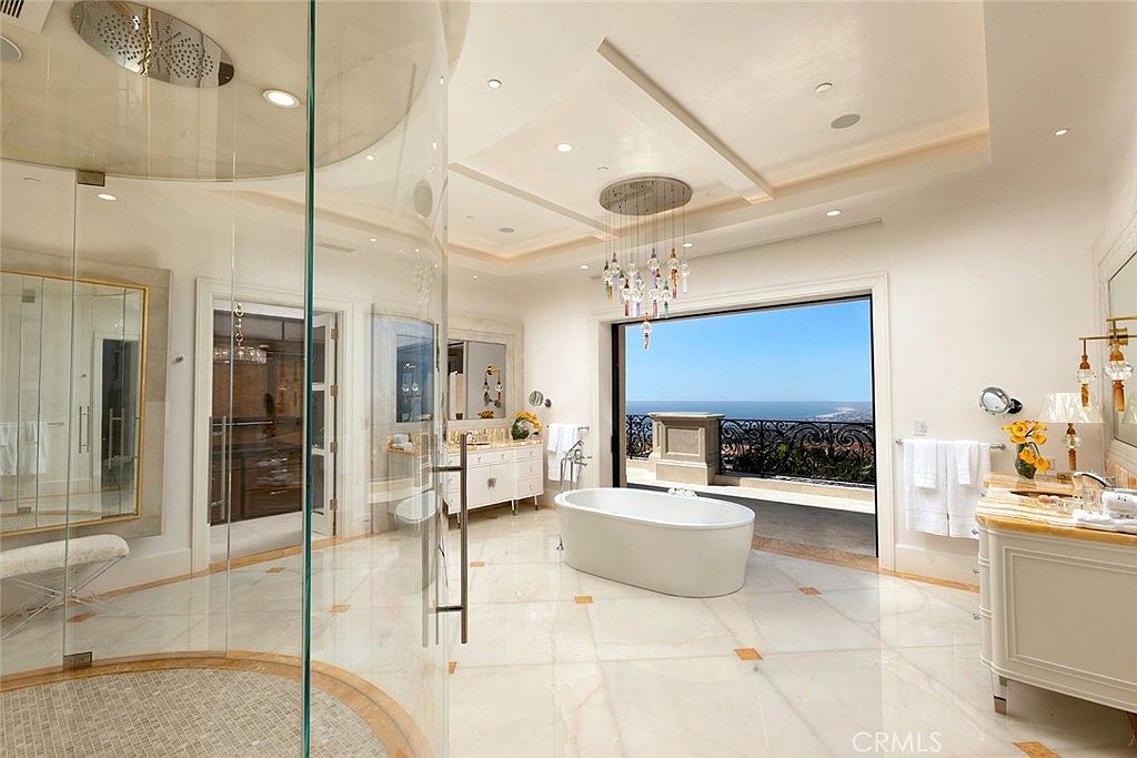 This is a luxurious primary bathroom featuring a glass-enclosed shower, a freestanding bathtub positioned to take advantage of the ocean view, and elegant white vanities with marble countertops. The room is spacious and well-lit, with a chandelier adding a touch of glamour. The large window/door provides access to a balcony, enhancing the spa-like atmosphere.