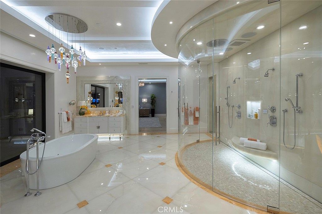 This is a luxurious primary bathroom featuring a freestanding bathtub with chrome fixtures, a spacious glass-enclosed shower with multiple showerheads and a built-in bench, and a vanity with marble countertops. The flooring is marble tile with decorative inlays, and a modern chandelier hangs above the tub. The overall impression is elegant and spa-like.