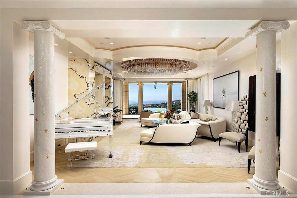 This is a luxurious living room featuring a grand piano, elegant seating, and a stunning chandelier. The room boasts marble columns, a view of the ocean, and a sophisticated color palette, creating an opulent and inviting atmosphere. The perspective is from the entrance of the room.