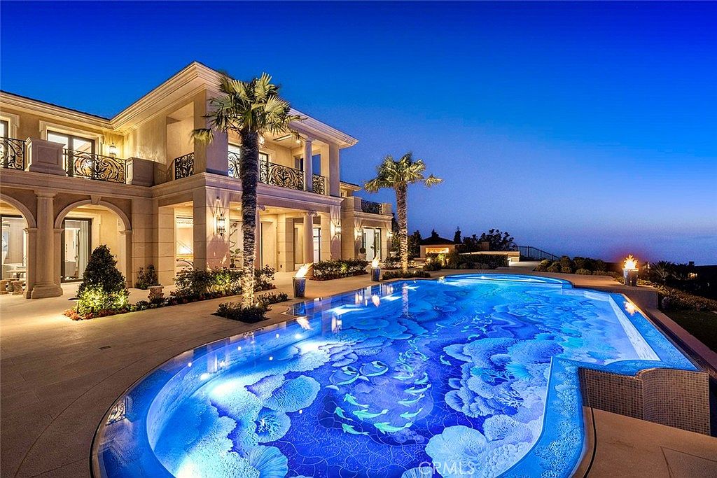 This image showcases a luxurious outdoor pool area at dusk, featuring a large, uniquely designed pool with intricate tile work. The pool is surrounded by a well-manicured patio and lush landscaping, with a grand two-story house in the background, complete with balconies and architectural details. The scene exudes opulence and relaxation, perfect for high-end real estate appeal.