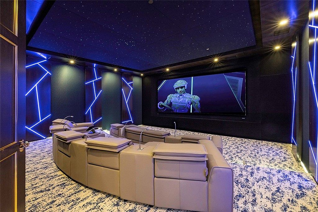 This is a high-end home theater room featuring a large curved sectional sofa with individual headrests, facing a large screen displaying a scene from Tron. The room is decorated with blue neon geometric designs on the walls and a starry night ceiling, creating a futuristic and immersive viewing experience. The carpet has a modern, abstract pattern.