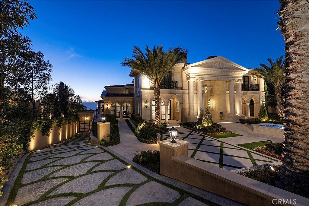 This image showcases the grand front exterior of a luxurious estate at dusk. The architecture features prominent columns, a stately entrance, and meticulously landscaped grounds with patterned driveway and mature palm trees. Warm lighting accentuates the architectural details, creating an inviting and impressive curb appeal.