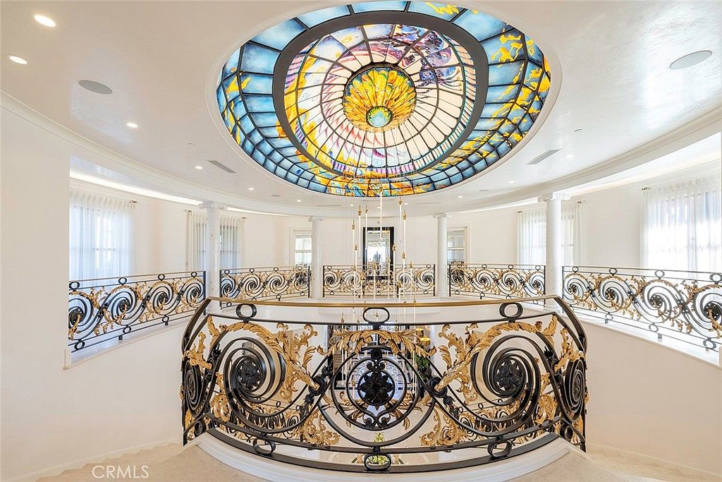 This interior shot showcases a grand hallway with ornate wrought iron railings accented with gold details, leading to a staircase. A stunning stained-glass dome ceiling provides a focal point, bathing the space in colorful light. The overall impression is one of luxury and architectural sophistication.