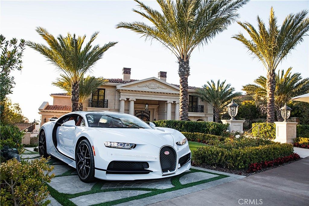This image showcases the front exterior of a luxurious property, featuring a white Bugatti Chiron parked on a paved driveway. The house boasts a grand entrance with columns, a tiled roof, and manicured landscaping including palm trees and hedges. The overall impression is one of opulence and high-end living.