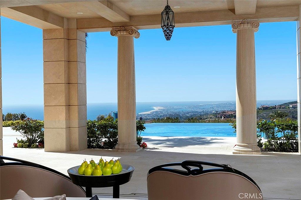 This image showcases a luxurious patio or balcony with grand architectural columns framing a stunning ocean view. The space features elegant outdoor furniture and a decorative hanging lantern, creating an inviting atmosphere. An infinity pool blends seamlessly with the horizon, enhancing the property's appeal as a high-end coastal retreat.