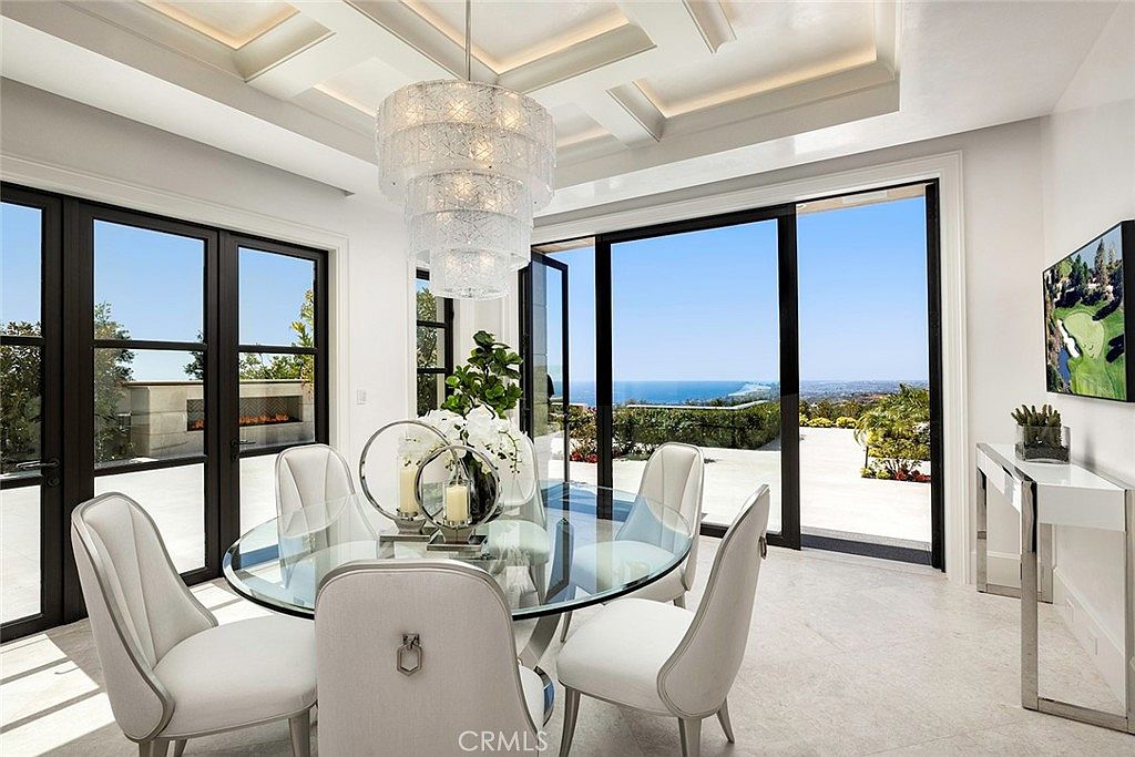 This is an interior shot of a dining room featuring a round glass table with six white upholstered chairs. A modern chandelier hangs above the table, and large sliding glass doors offer a view of the ocean. The room is bright and airy, with a clean, contemporary design.