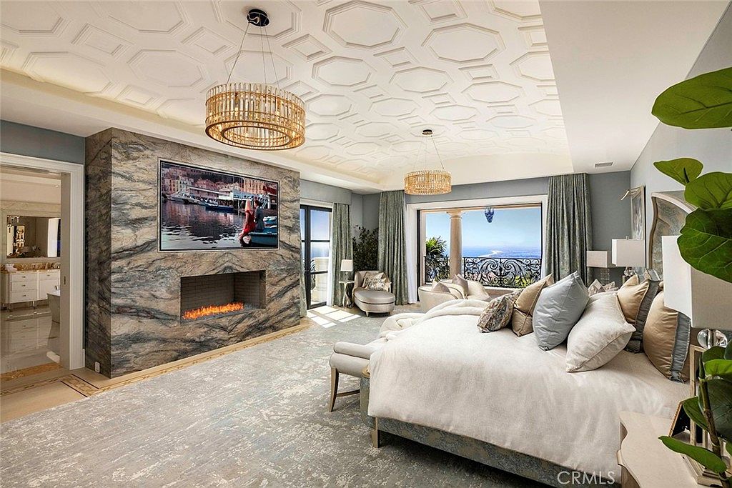 This is a luxurious primary bedroom featuring a large bed with plush pillows, a fireplace with a TV mounted above, and access to a balcony with ocean views. The room is decorated in a neutral color palette with elegant lighting fixtures and a textured ceiling, creating a serene and upscale atmosphere. The room has a wide angle perspective to show the entire room.