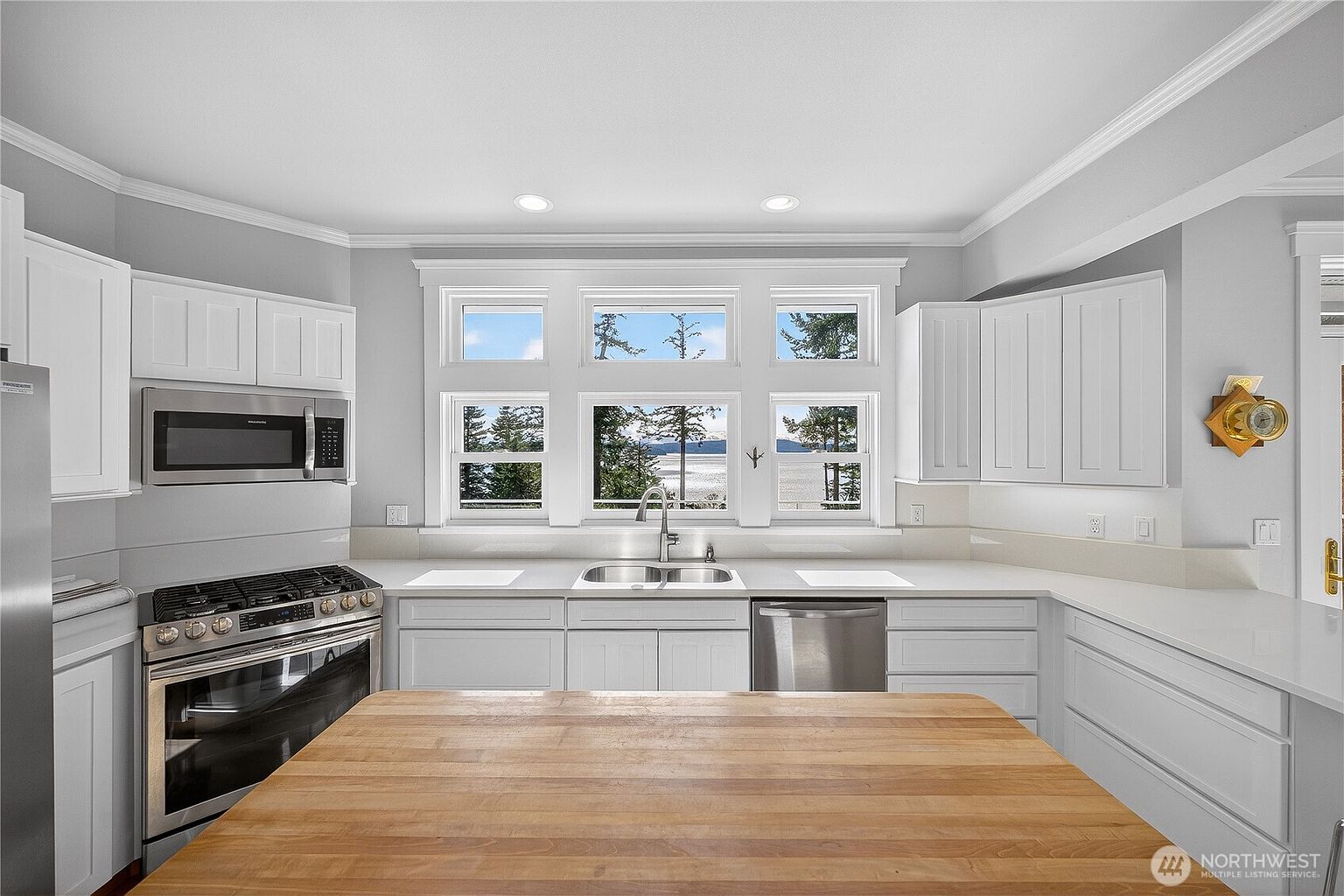 This bright and airy kitchen features crisp white cabinetry, a butcher block island, and high-end stainless steel appliances, including a gas range and built-in microwave. A large, multi-pane window above the sink provides a stunning, panoramic view of the water and surrounding trees, flooding the space with natural light. The clean, minimalist aesthetic is complemented by white countertops and a neutral color palette, creating a serene and inviting atmosphere.