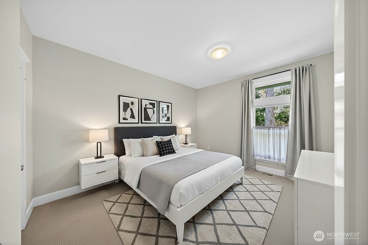This bright and airy bedroom features a neutral color palette with a modern, minimalist aesthetic. The room is centered around a queen-sized bed with a dark upholstered headboard, flanked by two white nightstands and matching lamps. A large window with floor-to-ceiling curtains allows for ample natural light, while a geometric-patterned area rug adds texture to the carpeted floor.