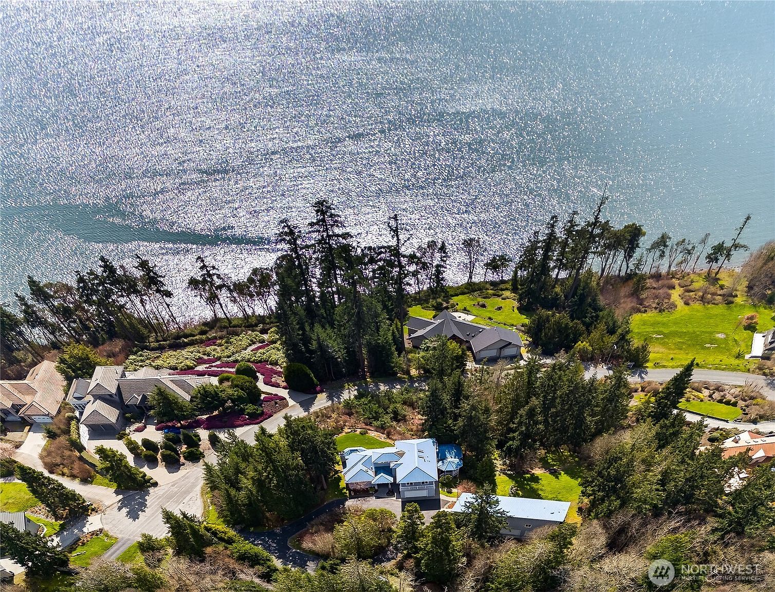 This high-angle aerial view captures a scenic coastal property featuring multiple luxury homes nestled among lush, mature evergreen trees. The landscape is characterized by winding driveways, vibrant garden beds, and expansive green lawns that lead toward a sparkling, sun-drenched body of water. The perspective provides a comprehensive look at the estate's layout and its prime waterfront location.