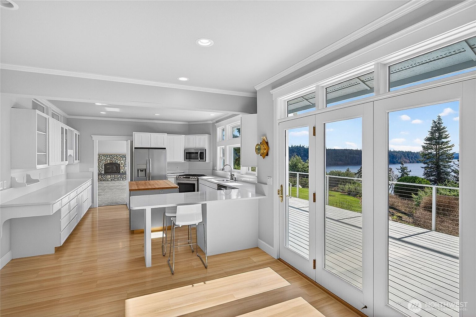 This bright and airy kitchen features crisp white cabinetry, sleek quartz countertops, and a central island with a butcher block top. The space flows seamlessly into an outdoor deck through large glass French doors, which provide expansive views of the surrounding landscape and water. The open-concept design, combined with light wood flooring and recessed lighting, creates a clean, modern, and inviting atmosphere.