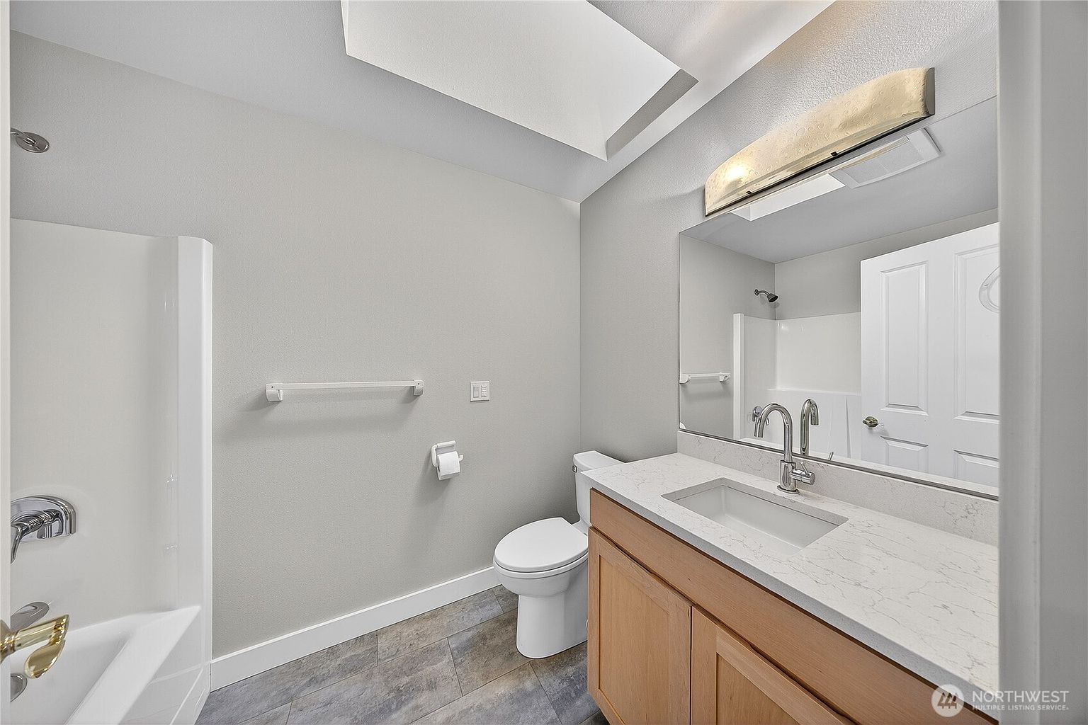 This clean and modern bathroom features a light wood vanity topped with a white quartz countertop and a rectangular undermount sink. The space includes a neutral gray color palette, a large wall-mounted mirror, and a combination shower-tub unit with white surround tiles. The room is well-lit by a sleek vanity light fixture and a skylight, creating a bright and inviting atmosphere.