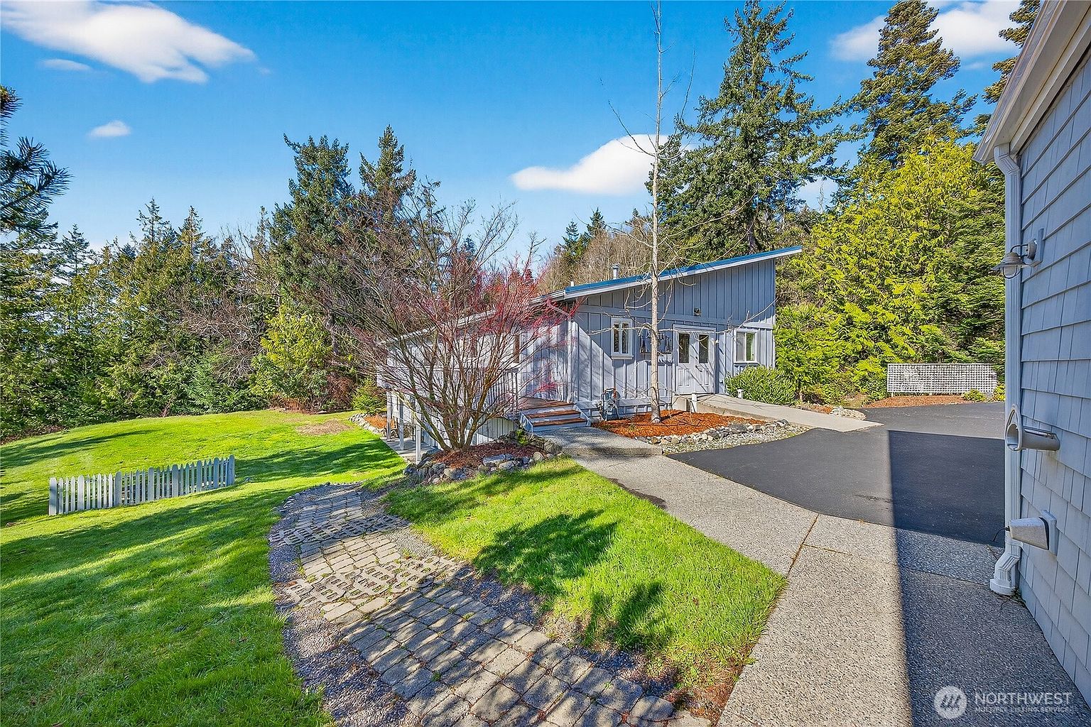 This exterior view captures a charming, light-blue wood-sided home with a distinctive shed-style roofline, nestled amidst a lush, tree-filled landscape. A stone-paved walkway leads from the foreground through a vibrant green lawn toward the home's entryway, which features a small wooden deck and double doors. The scene is bathed in bright, natural daylight, highlighting the peaceful, wooded setting and the home's inviting curb appeal.