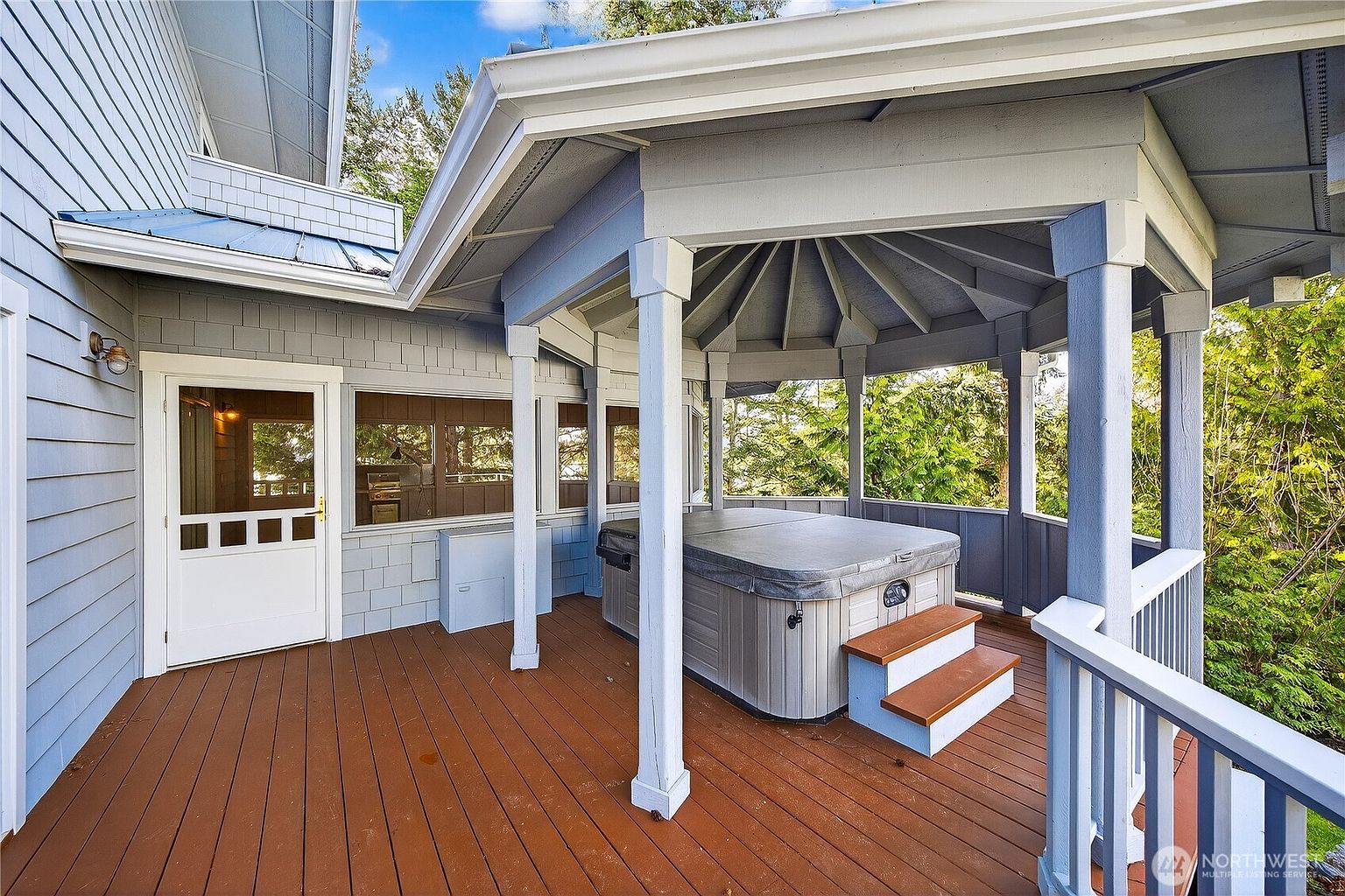 This inviting outdoor deck features a covered gazebo area housing a hot tub, complete with wooden steps for easy access. The space is finished with warm-toned decking and light blue siding, offering a serene and private atmosphere overlooking the surrounding trees. The perspective captures the transition from the home's interior access point to the relaxing spa retreat.