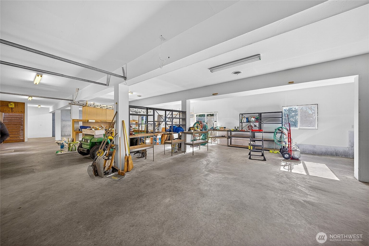 This spacious, unfinished garage features a vast concrete floor and an open-concept layout supported by structural pillars. The area is equipped with industrial-style metal shelving units, various gardening tools, and a small riding lawn mower, suggesting ample storage and workspace potential. The perspective is wide-angle, capturing the depth and functional utility of this large, multi-purpose storage area.