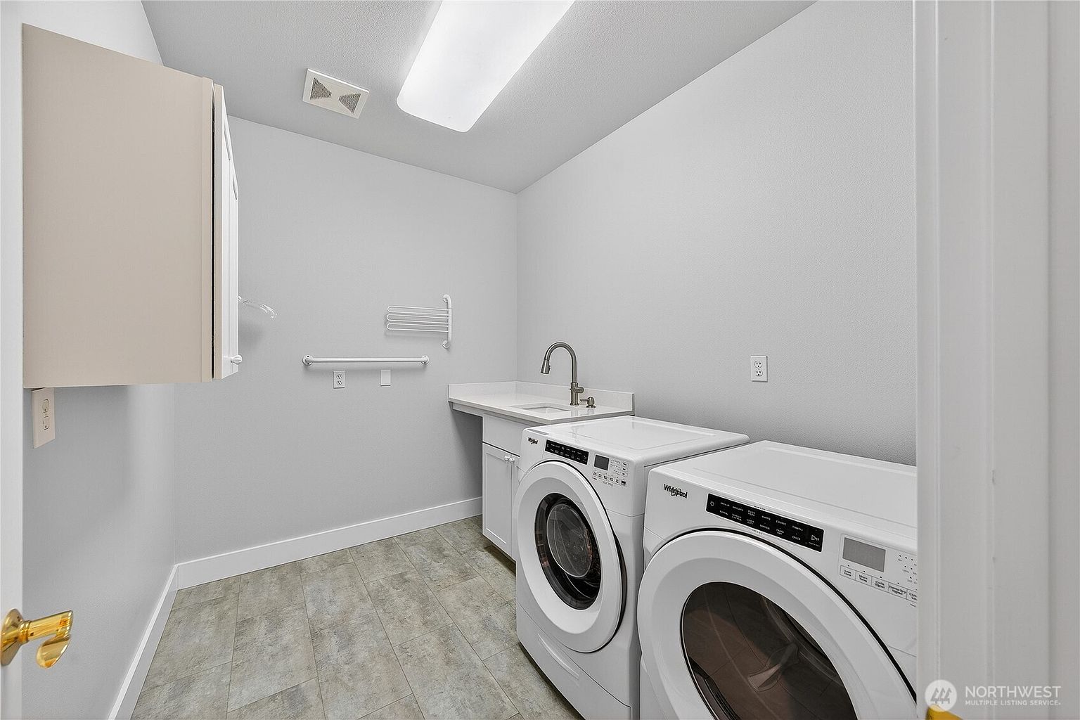 This clean and functional laundry room features white cabinetry, a utility sink with a modern faucet, and a pair of side-by-side Whirlpool washer and dryer units. The space is finished with light grey walls and durable, neutral-toned flooring, providing a bright and organized atmosphere. A wall-mounted drying rack and upper storage cabinet add practical utility to this well-maintained area.
