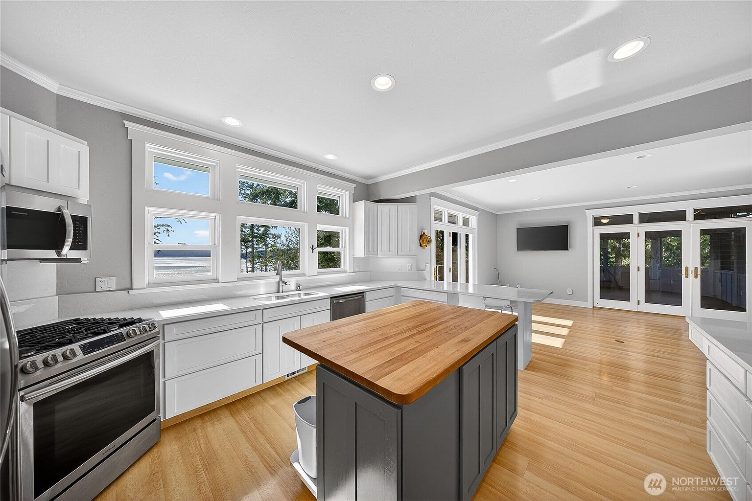 This bright and airy kitchen features a central island with a warm wood countertop, contrasting beautifully against the crisp white cabinetry and light hardwood flooring. The space is illuminated by recessed lighting and large windows that offer a scenic view of the water, creating an inviting and open atmosphere. The layout flows seamlessly into an adjacent living area, making it an ideal space for both cooking and entertaining.