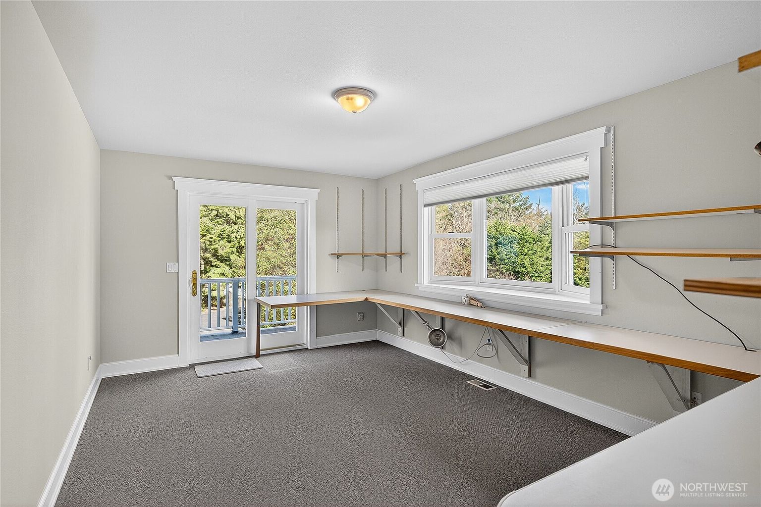 This bright and functional home office features a large, L-shaped built-in desk spanning two walls, providing ample workspace. The room is illuminated by a wide window and a glass-paned door that leads out to a balcony, offering a pleasant view of the surrounding greenery. With neutral-toned walls and grey carpeting, the space offers a clean, versatile environment suitable for a productive work-from-home setup.
