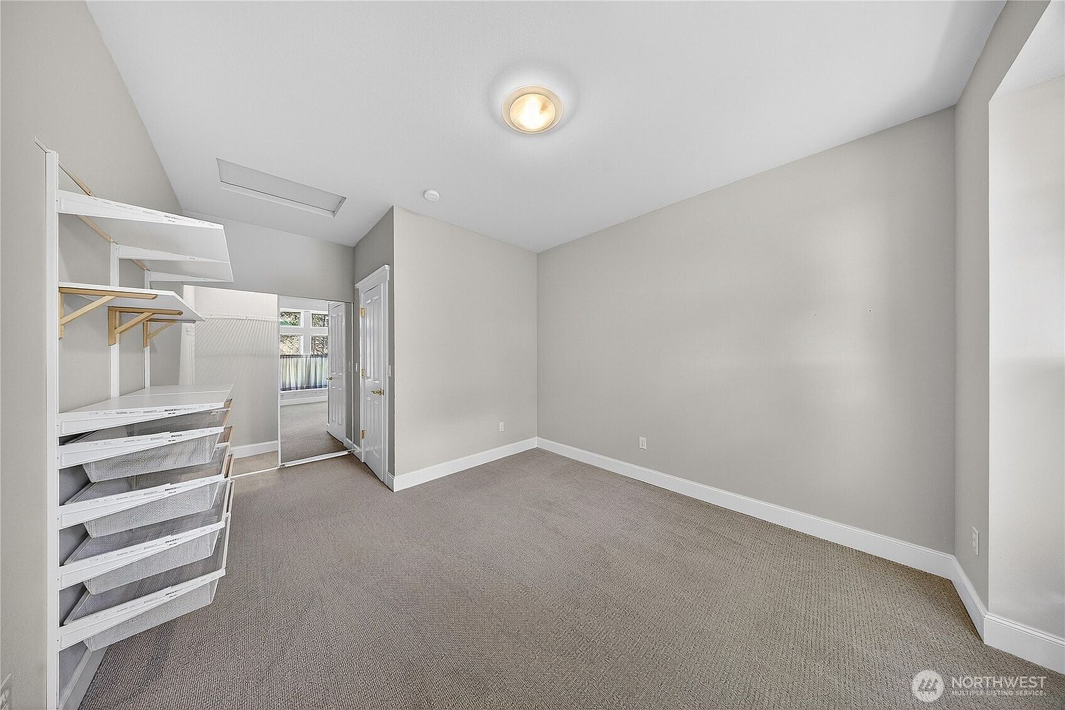 This image showcases a spacious walk-in closet featuring a neutral color palette with light gray walls and beige carpeting. The room is equipped with an extensive white wire shelving system, including multiple pull-out baskets and hanging rods for organized storage. A doorway leads into an adjacent room, providing a sense of depth and flow within the home's layout.