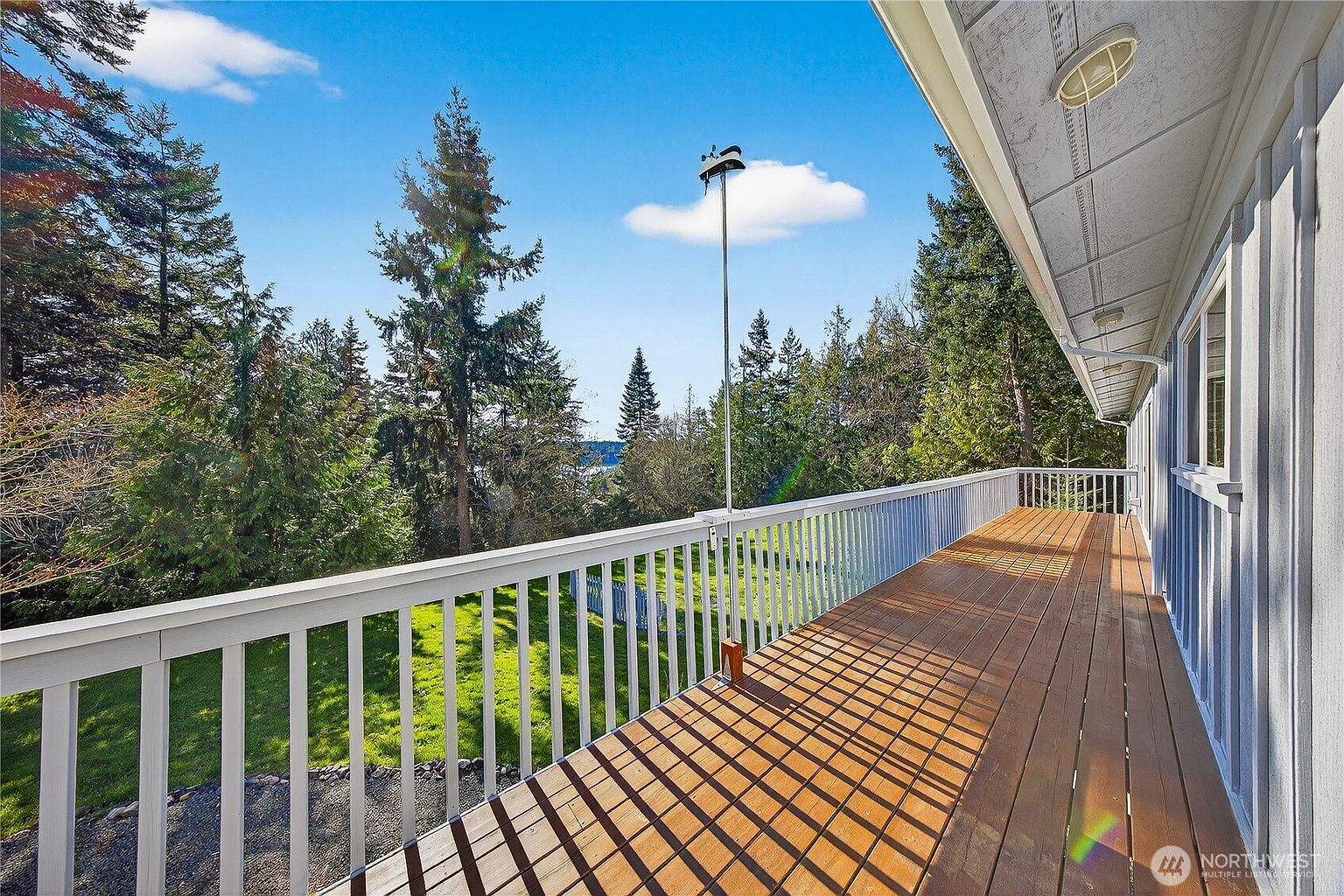 This image showcases a spacious, elevated wooden deck that runs along the side of a house, featuring a white railing and a clear view of the surrounding lush greenery and distant water. The perspective is from the deck looking outward, capturing the warm sunlight casting long shadows across the wooden planks. It offers a serene and inviting outdoor living space perfect for relaxation or entertaining while enjoying the natural scenery.