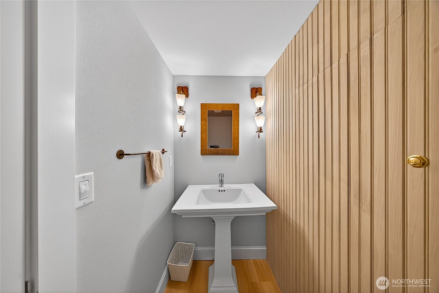 This compact powder room features a classic pedestal sink centered against a light gray wall, flanked by two sets of dual-bulb wall sconces. To the right, a striking floor-to-ceiling vertical wood slat accent wall adds warmth and texture to the space. The room is finished with light hardwood flooring and a small, framed mirror, creating a clean and modern aesthetic.