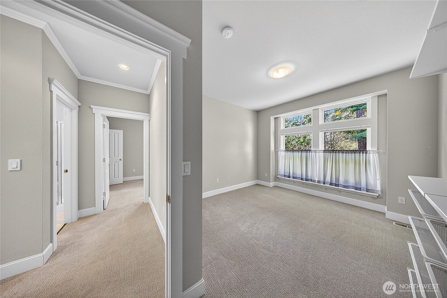 This interior view captures a bright, neutral-toned bedroom featuring plush carpeting and a large window that allows for ample natural light. The perspective is from a hallway looking into the room, showcasing the clean architectural lines, white trim, and a seamless transition between spaces. The room offers a versatile, blank-canvas aesthetic suitable for a guest bedroom or home office.