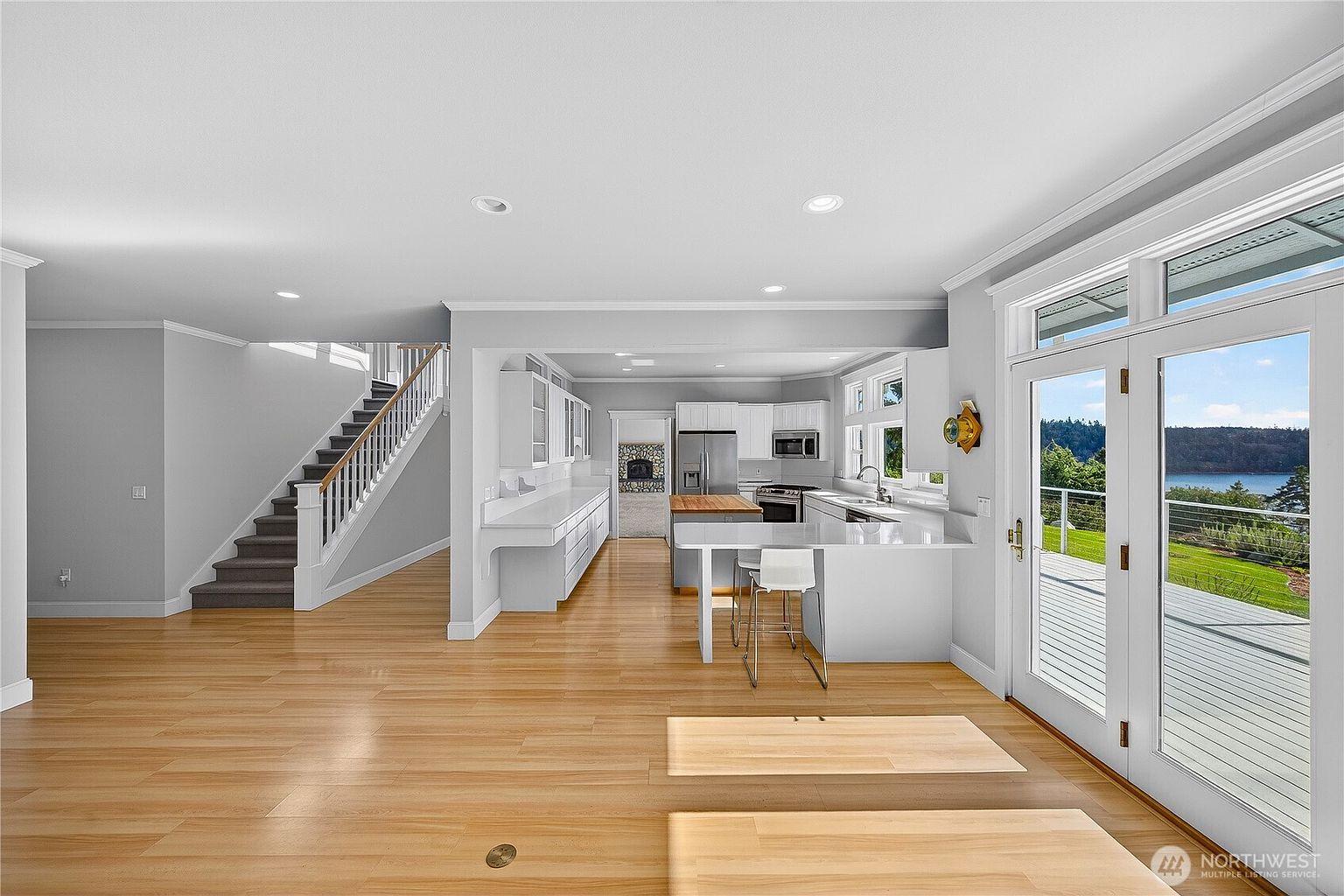 This bright, open-concept kitchen features sleek white cabinetry, a central island with a wooden butcher block top, and a breakfast bar with modern stools. The space flows seamlessly into a living area with a staircase, while large glass doors provide a stunning view of the outdoor deck and water beyond. The light hardwood flooring and recessed lighting create a clean, airy, and inviting atmosphere.