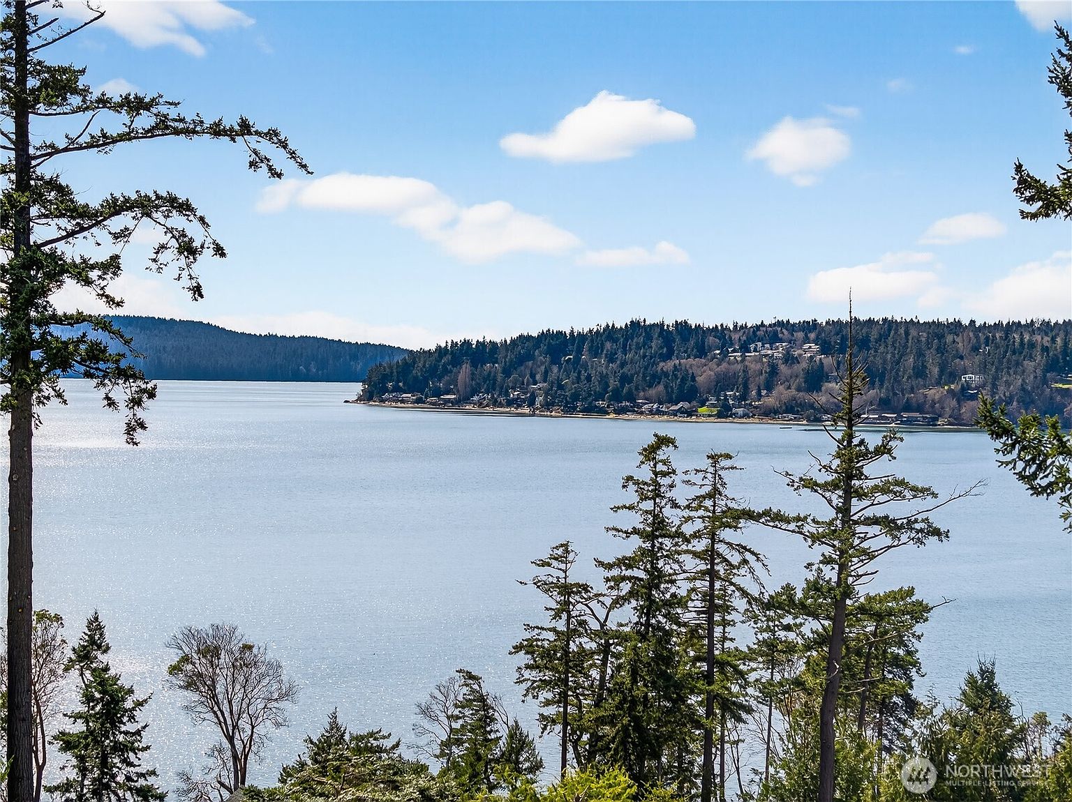 This image captures a scenic, elevated view of a calm body of water, likely a bay or sound, framed by evergreen trees in the foreground. The perspective is from a high vantage point, such as a balcony or deck, looking out toward a distant, forested shoreline dotted with residential homes. The bright, clear sky and expansive water create a serene and picturesque atmosphere, highlighting the property's desirable waterfront location.