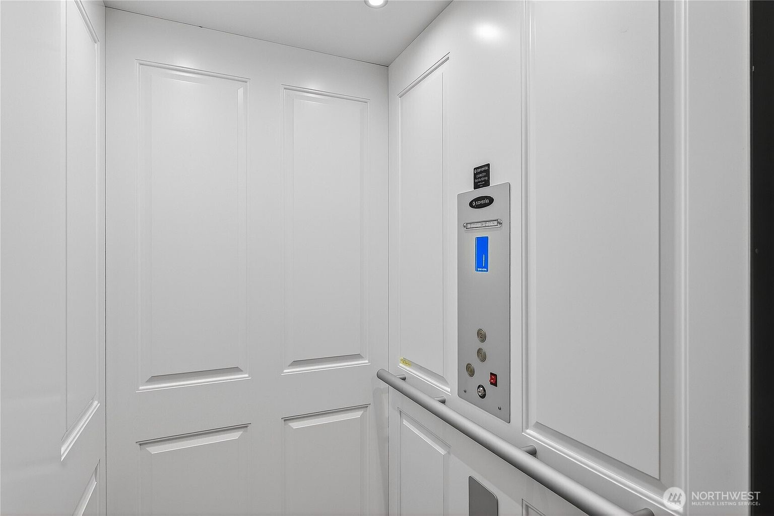 This image shows the interior of a residential elevator, featuring clean white paneled walls and a sleek, modern control panel. The elevator is equipped with a metal handrail and a digital display indicating floor levels, reflecting a high-end, accessible home feature. The perspective is a straight-on view from inside the small, enclosed space, emphasizing its functional and polished design.