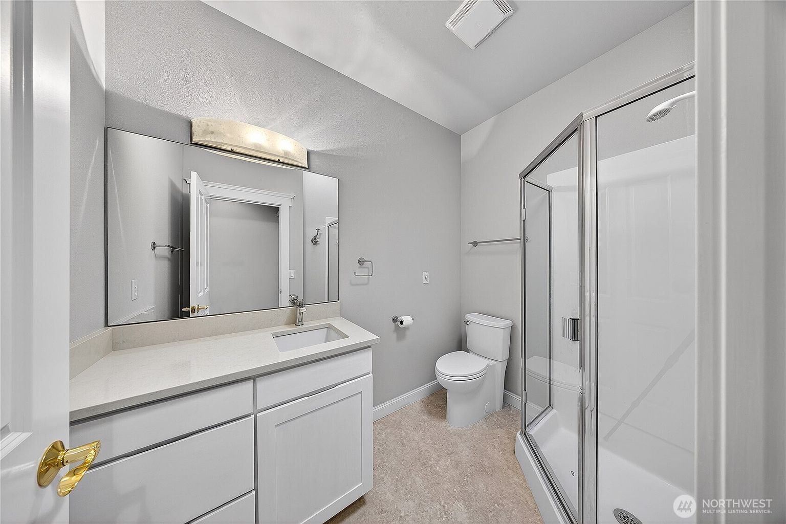 This clean and modern bathroom features a white vanity with a quartz countertop, a large wall-mounted mirror, and a glass-enclosed walk-in shower. The space is finished with neutral grey walls and light-colored flooring, creating a bright and functional atmosphere. The perspective is from the doorway, capturing the entire layout including the toilet and vanity area.