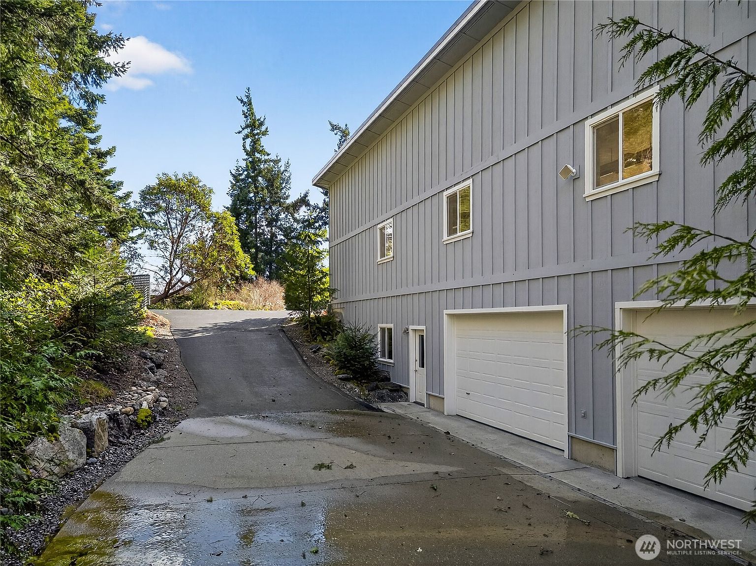 This side view captures a multi-level home with light gray vertical siding and a paved driveway leading up to a two-car garage. The perspective highlights the transition from the concrete driveway to the asphalt slope, framed by lush evergreen trees and natural landscaping. The scene conveys a quiet, suburban atmosphere with a focus on the property's functional exterior layout.