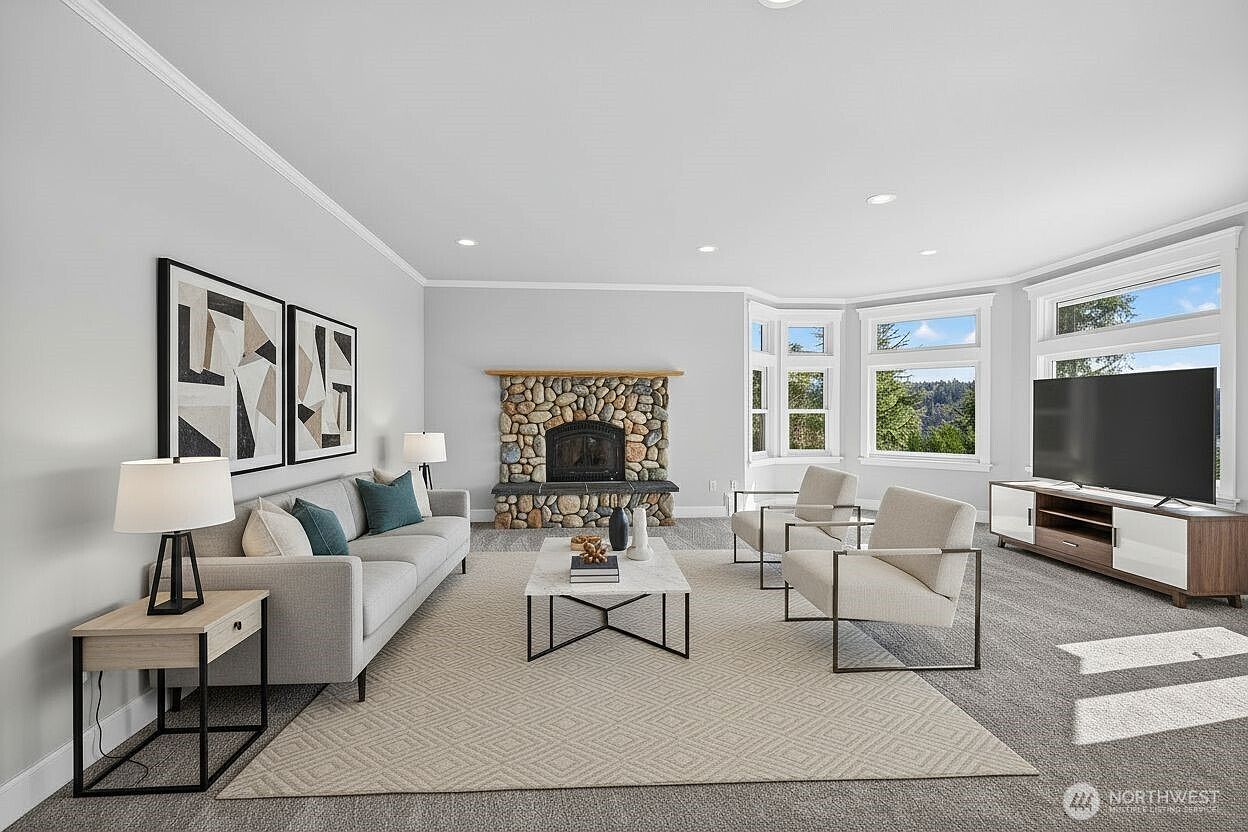 This bright and spacious living room features a neutral color palette, plush carpeting, and a striking stone fireplace as the central focal point. The room is furnished with a modern grey sofa, two accent chairs, and a sleek coffee table, all arranged on a large area rug. Large windows allow for ample natural light, creating an inviting and comfortable atmosphere perfect for relaxation.