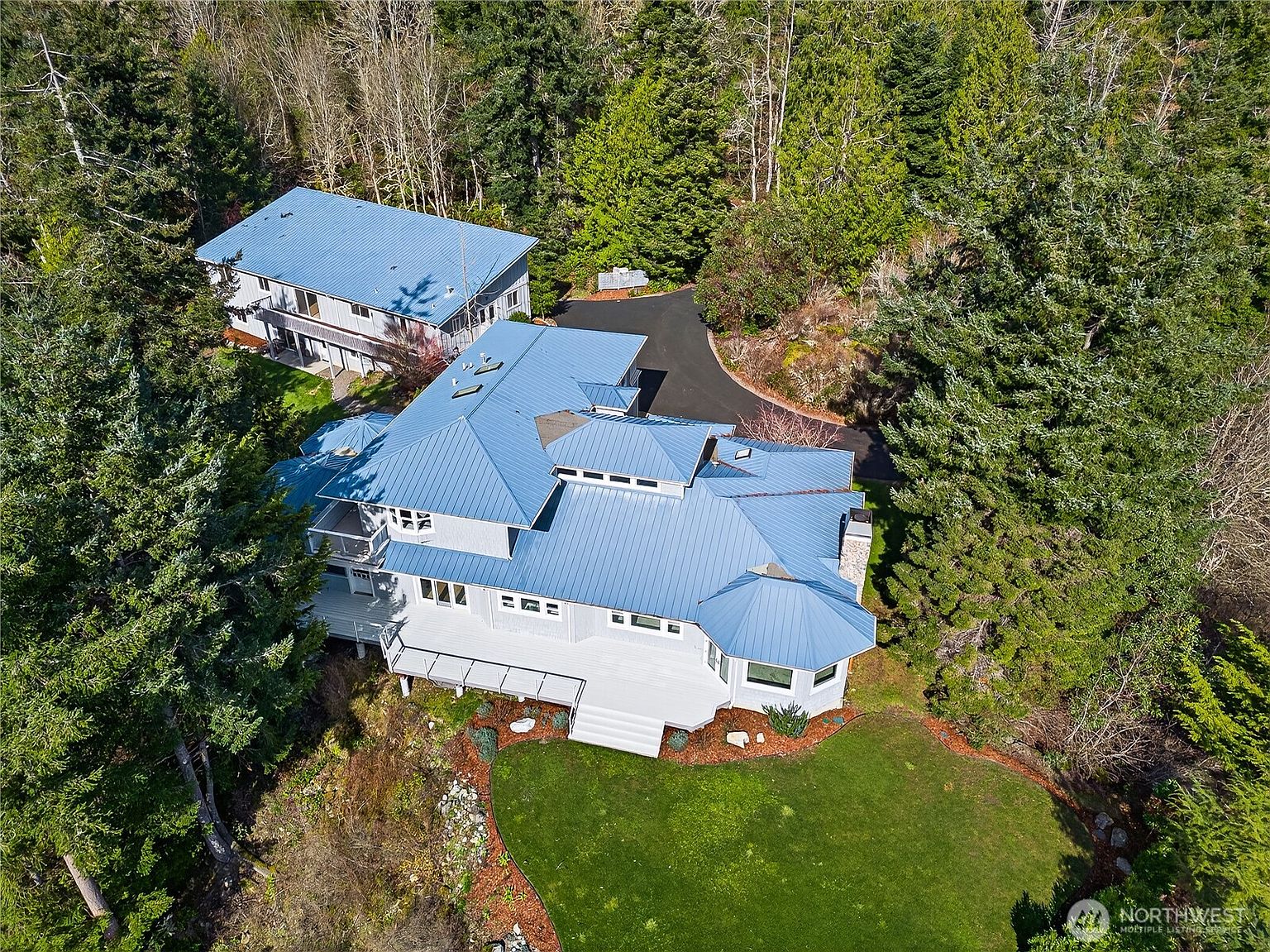 This high-angle aerial view captures a large, multi-level home with a distinctive blue metal roof nestled within a dense, lush forest. The property features an expansive white deck overlooking a manicured lawn, a secondary detached building, and a winding paved driveway. The setting offers a serene, secluded atmosphere, highlighting the home's integration with the surrounding natural landscape.