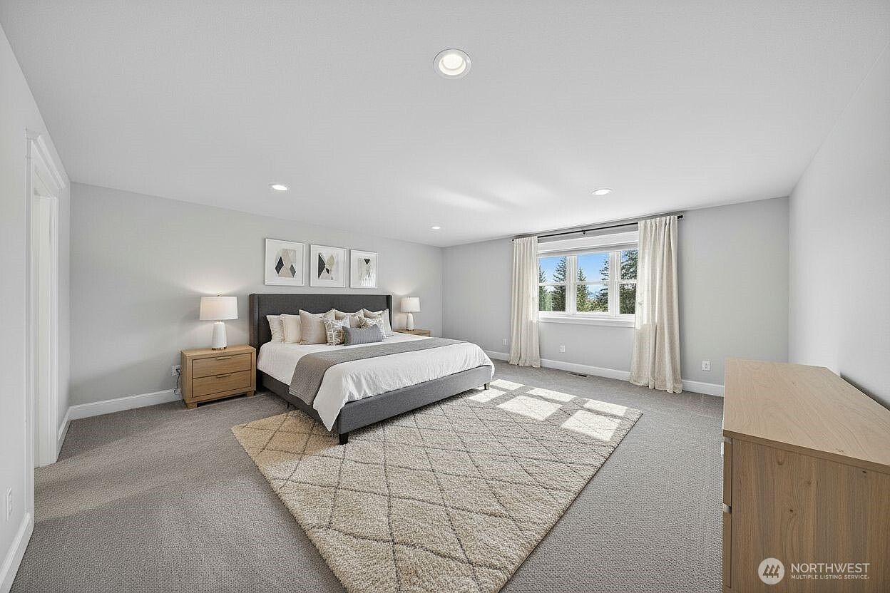 This spacious primary bedroom features a neutral color palette, plush carpeting, and a large, centered bed with a grey upholstered headboard. A geometric-patterned area rug anchors the room, while large windows with elegant curtains allow for ample natural light. The room is minimalist and serene, offering a clean, modern aesthetic suitable for a primary suite.
