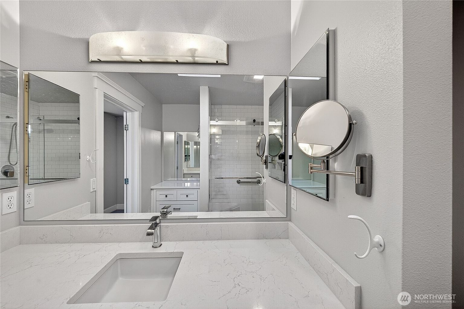 This modern bathroom features a clean, minimalist aesthetic with a white quartz vanity countertop and an undermount sink. A large, wall-to-wall mirror reflects the bright, neutral-toned space, including a glass-enclosed shower with white subway tile and a wall-mounted magnifying mirror. The perspective is a straight-on eye-level shot, emphasizing the bright, airy, and well-maintained condition of the room.