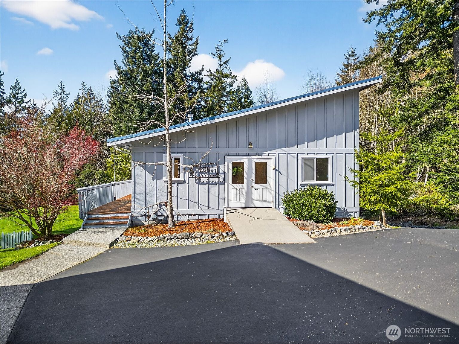 This exterior front view showcases a charming, single-story home with a distinctive shed-style roof and light blue vertical siding. The property features a paved driveway leading to a central double-door entryway with an accessible concrete ramp, complemented by a wooden deck on the left. Surrounded by lush evergreen trees and a well-maintained lawn, the home presents a peaceful, rustic aesthetic.