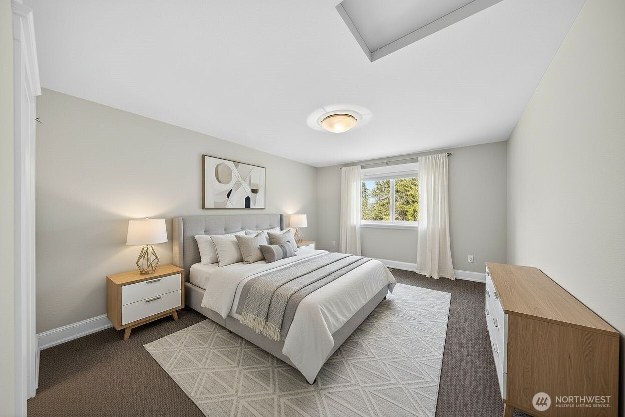 This bright and airy primary bedroom features a neutral color palette, a plush upholstered bed frame, and a matching wooden dresser. A large window with white curtains allows natural light to fill the room, while a geometric area rug adds texture to the dark carpeting. The space is styled with modern, minimalist decor, creating a serene and inviting atmosphere.