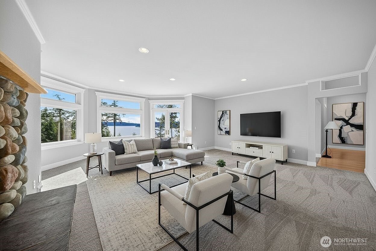 This spacious and bright living room features a neutral color palette, plush carpeting, and a large sectional sofa arranged around a modern coffee table. A stone-clad fireplace adds a rustic touch, while expansive windows provide a stunning view of the water, creating a serene and inviting atmosphere. The room is tastefully decorated with contemporary art and comfortable seating, making it an ideal space for relaxation and entertaining.