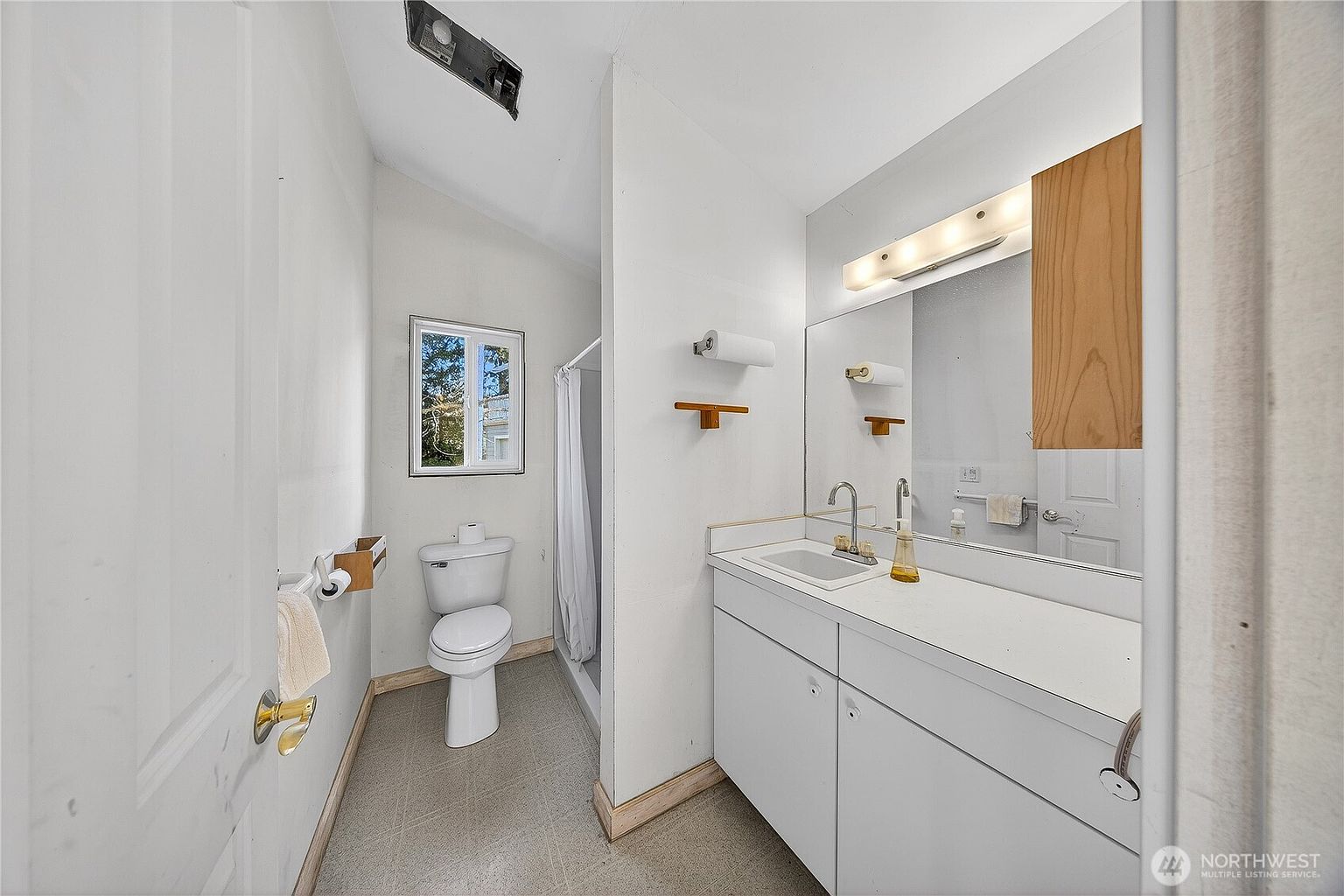 This functional bathroom features a white vanity with a single sink, a toilet, and a shower stall tucked behind a partition wall. The space is illuminated by a wall-mounted light fixture above a large mirror and includes a small window for natural light. The neutral color palette and simple layout create a clean, practical aesthetic suitable for a guest or secondary bathroom.
