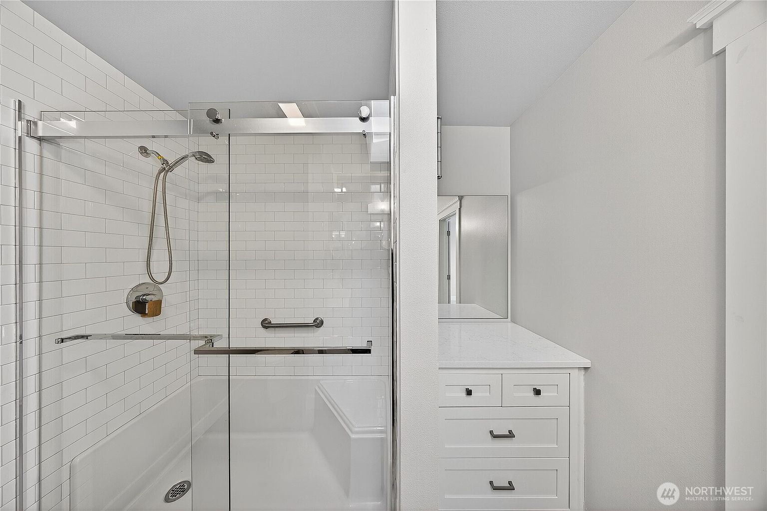 This modern bathroom features a sleek, walk-in glass shower with white subway tile walls and a built-in bench. Adjacent to the shower is a clean, white vanity with a quartz countertop and a mirror, creating a bright and minimalist aesthetic. The perspective captures the functional layout of the space, emphasizing the high-quality finishes and contemporary design.