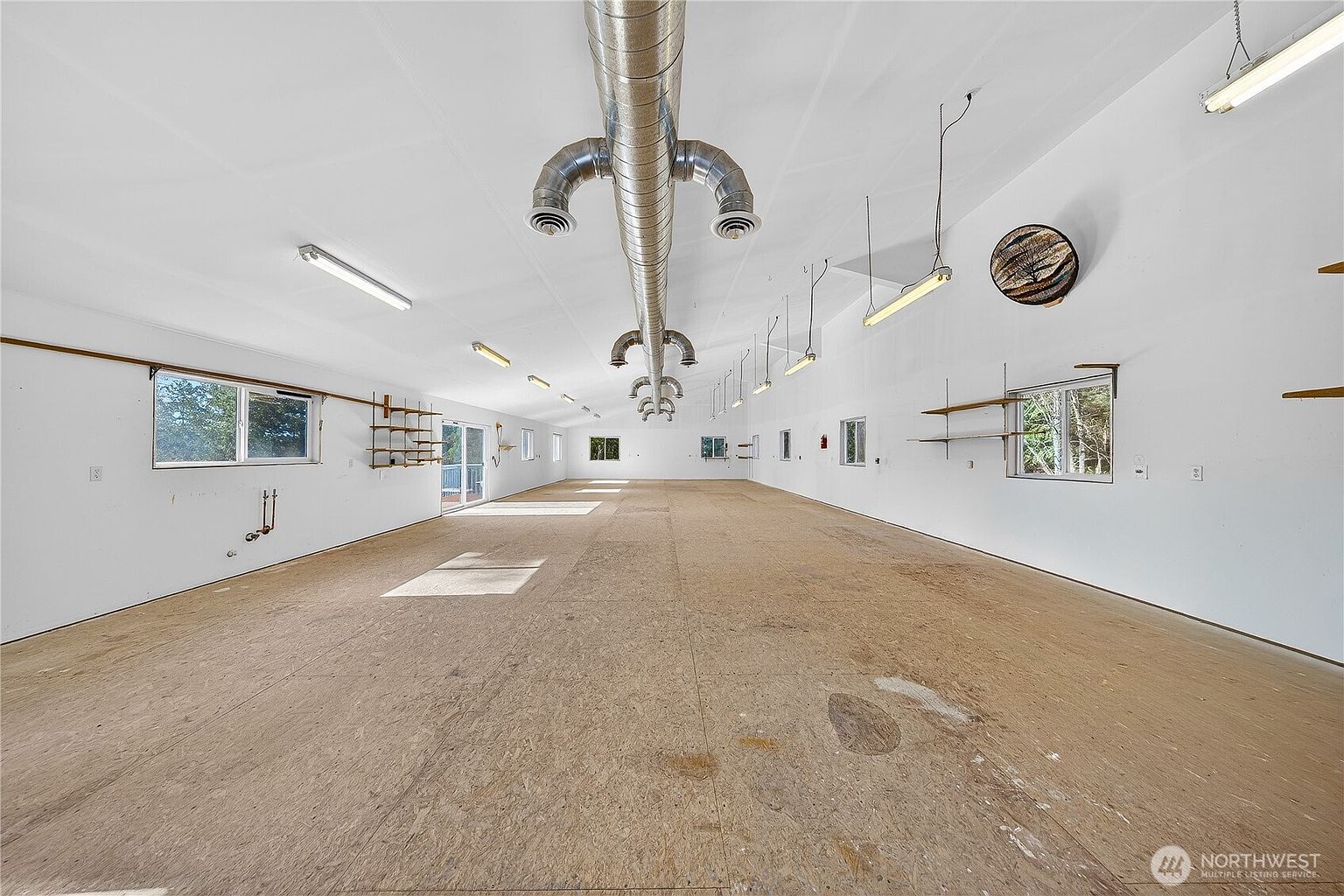 This image shows a large, unfinished interior space, likely a workshop or a multi-purpose garage area. The room features a long, central HVAC duct system running across the ceiling, white walls, and a plain concrete floor. Multiple windows line the walls, and there are several wall-mounted shelves and light fixtures, suggesting a functional, industrial-style workspace.