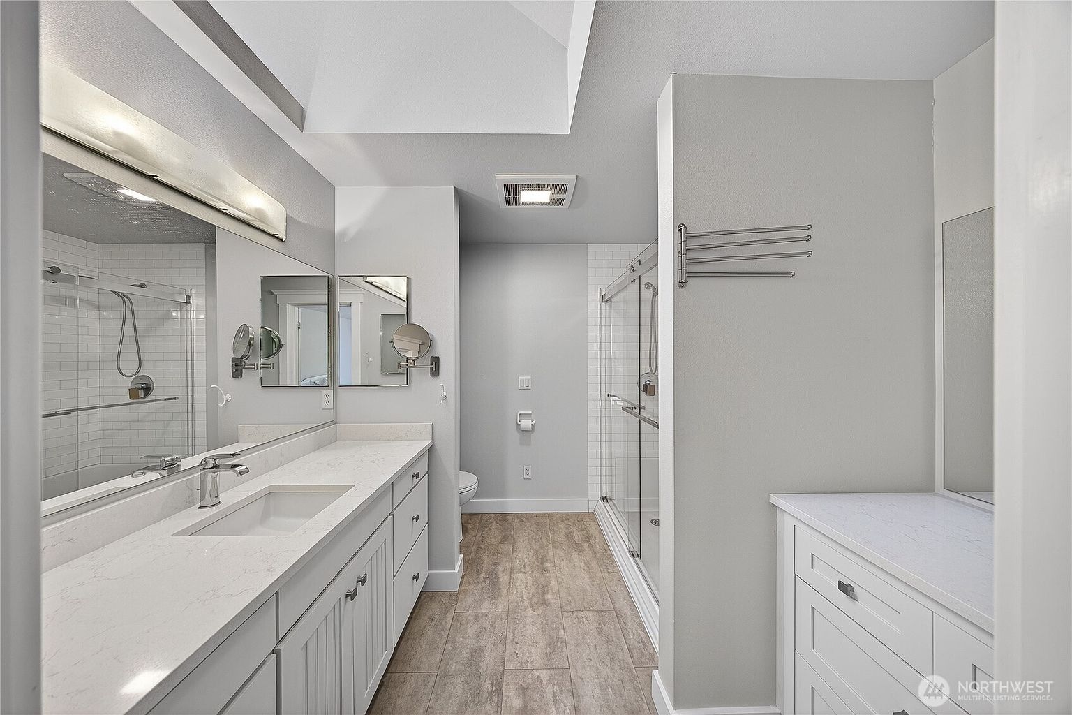 This modern primary bathroom features a long dual-sink vanity with white cabinetry and quartz countertops, complemented by a large wall-to-wall mirror. To the right, a glass-enclosed walk-in shower with subway tile accents adds a clean, contemporary aesthetic, while wood-look tile flooring runs throughout the space. The layout is bright and functional, leading toward a private toilet area at the back.