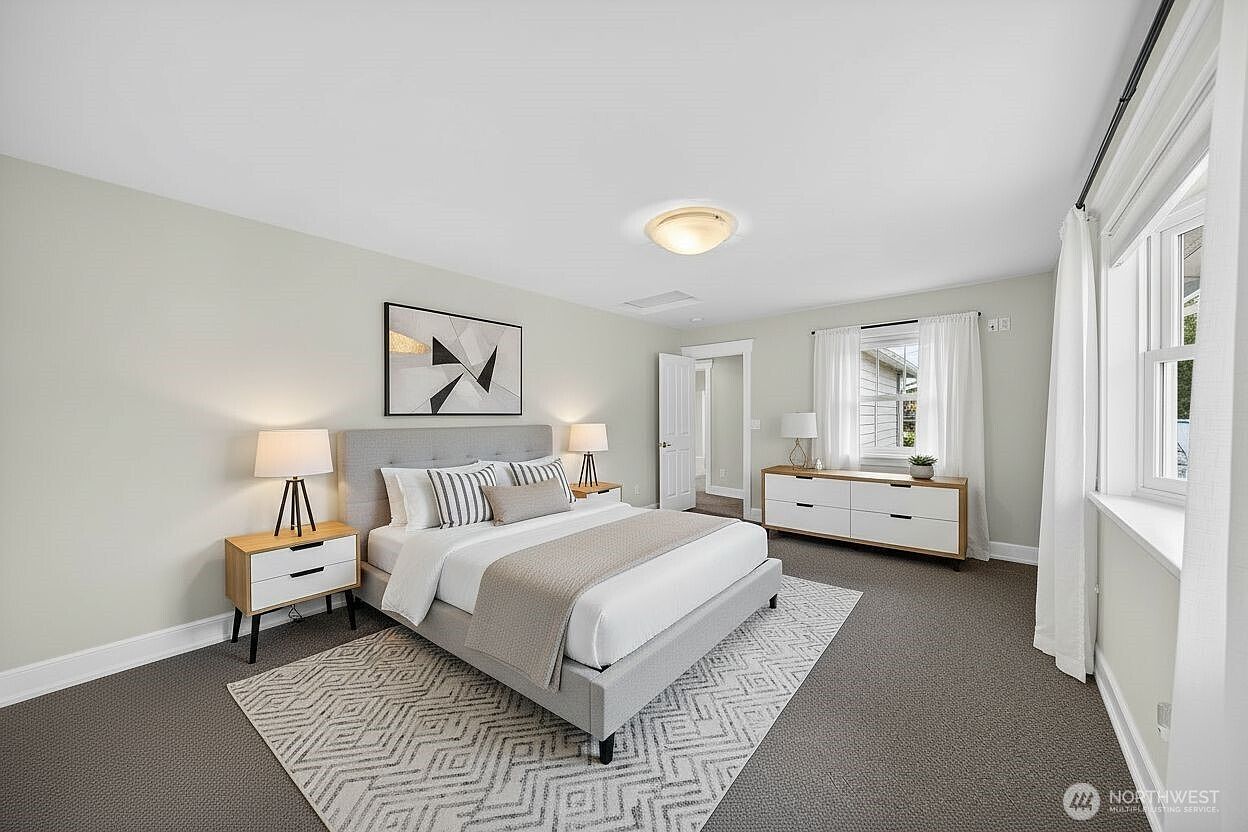 This spacious primary bedroom features a neutral color palette, plush carpeting, and a modern upholstered bed frame centered against a light gray wall. A geometric area rug adds texture to the room, while a white dresser and matching nightstands provide clean, contemporary storage. The perspective is a wide-angle shot from the corner, showcasing the bright, airy atmosphere created by the large windows and soft natural light.