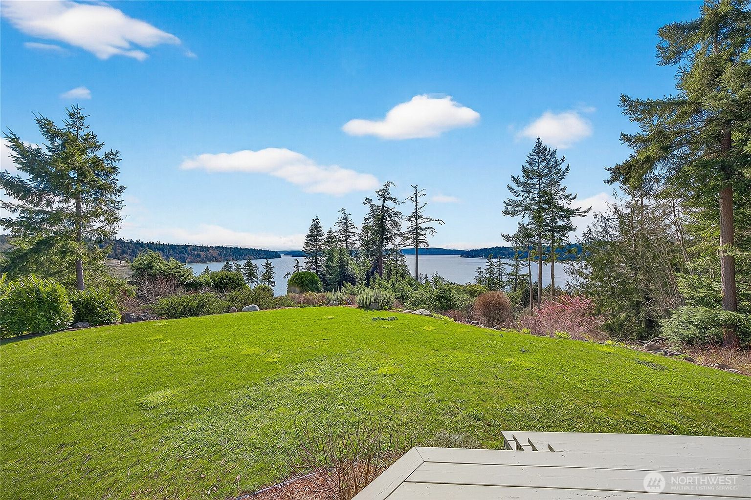 This image captures a serene, expansive backyard featuring a lush green lawn that leads toward a stunning, unobstructed view of a calm body of water and distant forested hills. The foreground includes a portion of a light-colored wooden deck, framing the picturesque landscape under a bright, partly cloudy sky. The scene evokes a sense of tranquility and privacy, highlighting the property's prime waterfront location.