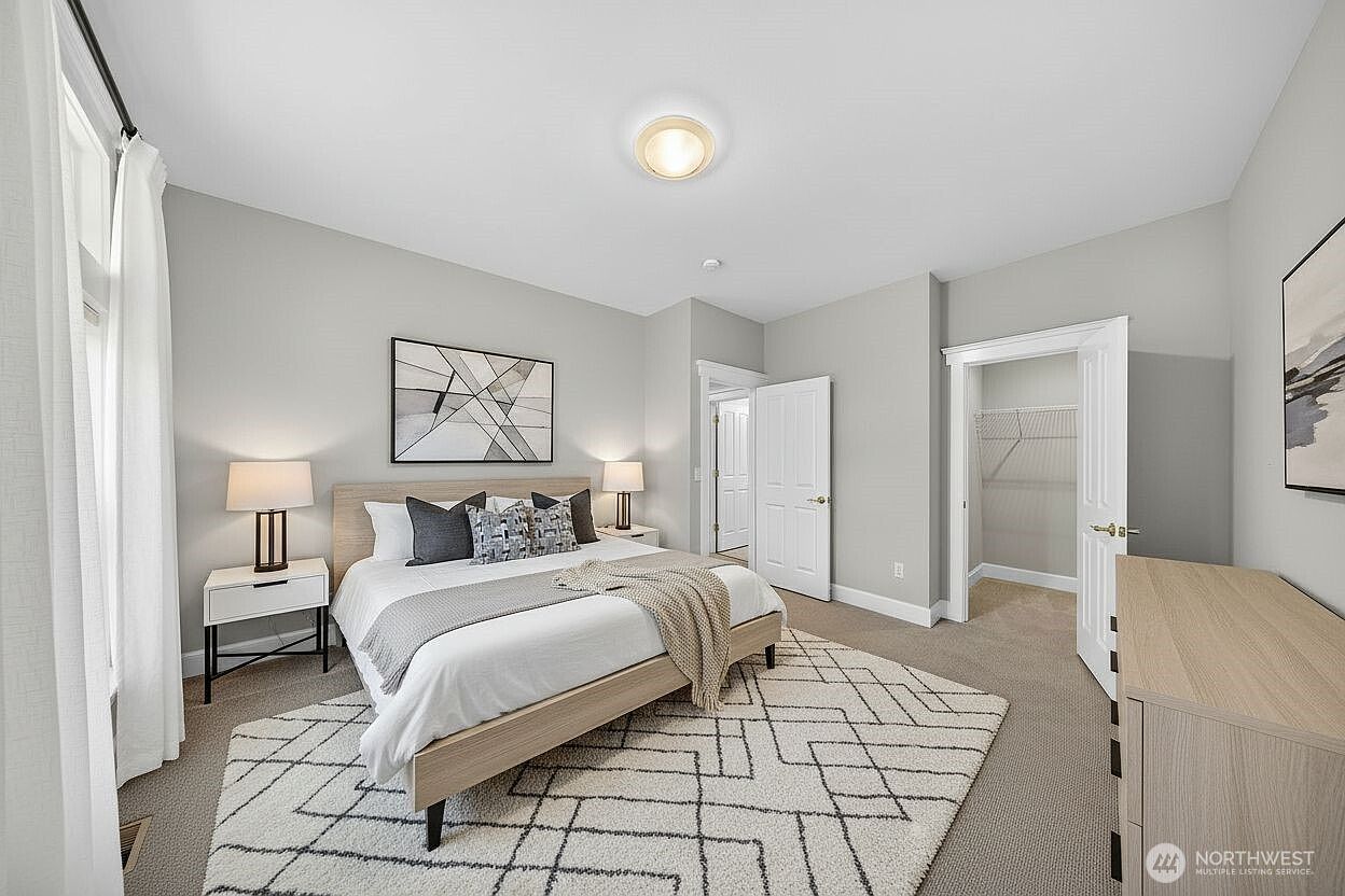 This bright and airy primary bedroom features a neutral color palette with soft gray walls and plush carpeting, creating a serene atmosphere. The room is anchored by a modern light-wood bed frame with a geometric-patterned rug, complemented by matching nightstands and lamps. A walk-in closet is visible through an open doorway, and the space is illuminated by a central ceiling light fixture, offering a clean and inviting aesthetic.