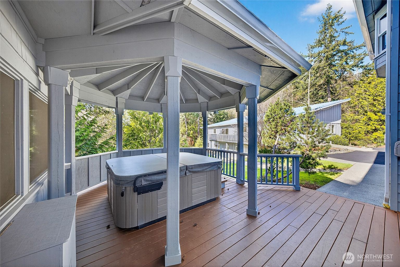 This image showcases a spacious wooden deck featuring a covered gazebo area that houses a large, grey hot tub. The deck is surrounded by a blue railing and offers a view of the surrounding trees and neighboring homes, creating a private and relaxing outdoor retreat. The perspective is from the deck looking toward the hot tub, emphasizing the functional layout and potential for outdoor leisure.
